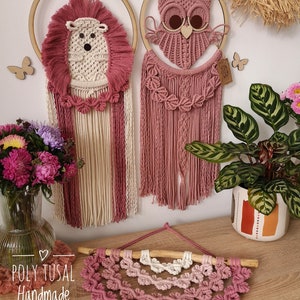 Pink Macrame Hedgehog and Warm Pink Macrame Owl hanging on the wall between the flowers. Pink verion of Macrame Leaf Half Mandala is laying on the table.
On the left is logo of Poly Tusal Handmade.