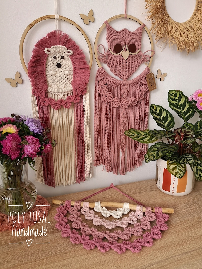 Pink Macrame Hedgehog and Warm Pink Macrame Owl hanging on the wall between the flowers. Pink verion of Macrame Leaf Half Mandala is laying on the table.
On the left is logo of Poly Tusal Handmade.