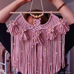 Pode incluir: Um pendurador de parede de macram&eacute; rosa feito &agrave; m&atilde;o, com uma vara de madeira e detalhes de contas. O design apresenta detalhes florais e de coruja, com franjas longas e contas de madeira. O texto "POLY TUSAL HANDMADE" &eacute; vis&iacute;vel.