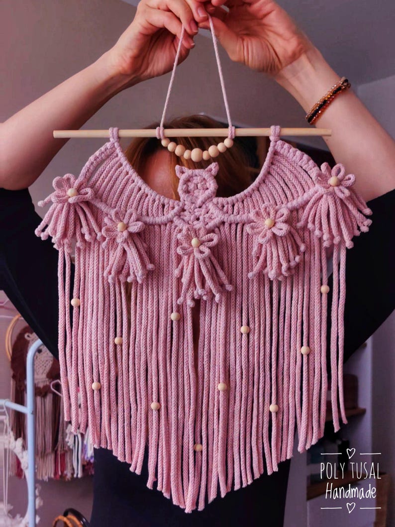 Pode incluir: Um pendurador de parede de macram&eacute; rosa feito &agrave; m&atilde;o, com uma vara de madeira e detalhes de contas. O design apresenta detalhes florais e de coruja, com franjas longas e contas de madeira. O texto "POLY TUSAL HANDMADE" &eacute; vis&iacute;vel.
