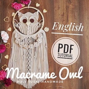 May include: A beige macrame owl wall hanging with a wooden hoop and fringe. The owl is made with a knotting technique and has large, round eyes. The background is a wooden surface with pink flowers and hearts.