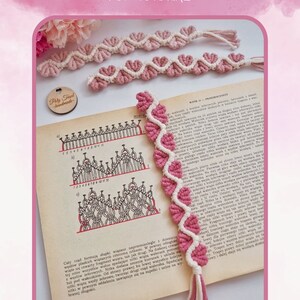 Op de afbeelding: Een roze en witte macram&eacute; hartvormige bladwijzer met een kwast. De bladwijzer is gemaakt met een knooptechniek genaamd macram&eacute;. De bladwijzer wordt getoond op een boek met een diagram van de knooptechniek.