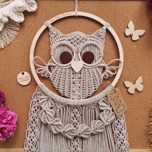 Op de afbeelding: Een handgemaakte macramé uil wanddecoratie met een houten frame en bruine accenten. De uil heeft ingewikkelde knoopdetails en is omgeven door decoratieve elementen, waaronder houten vlinders en bloemen. De tekst "Pily Tree Handmade" is zichtbaar.