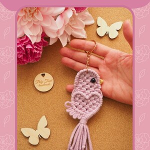 May include: A light purple macrame bird keychain with a heart-shaped body and tassel tail. The keychain has a gold ring and a black bead eye. The text "MACRAME BIRD KEYCHAIN" is visible. The design is handmade.