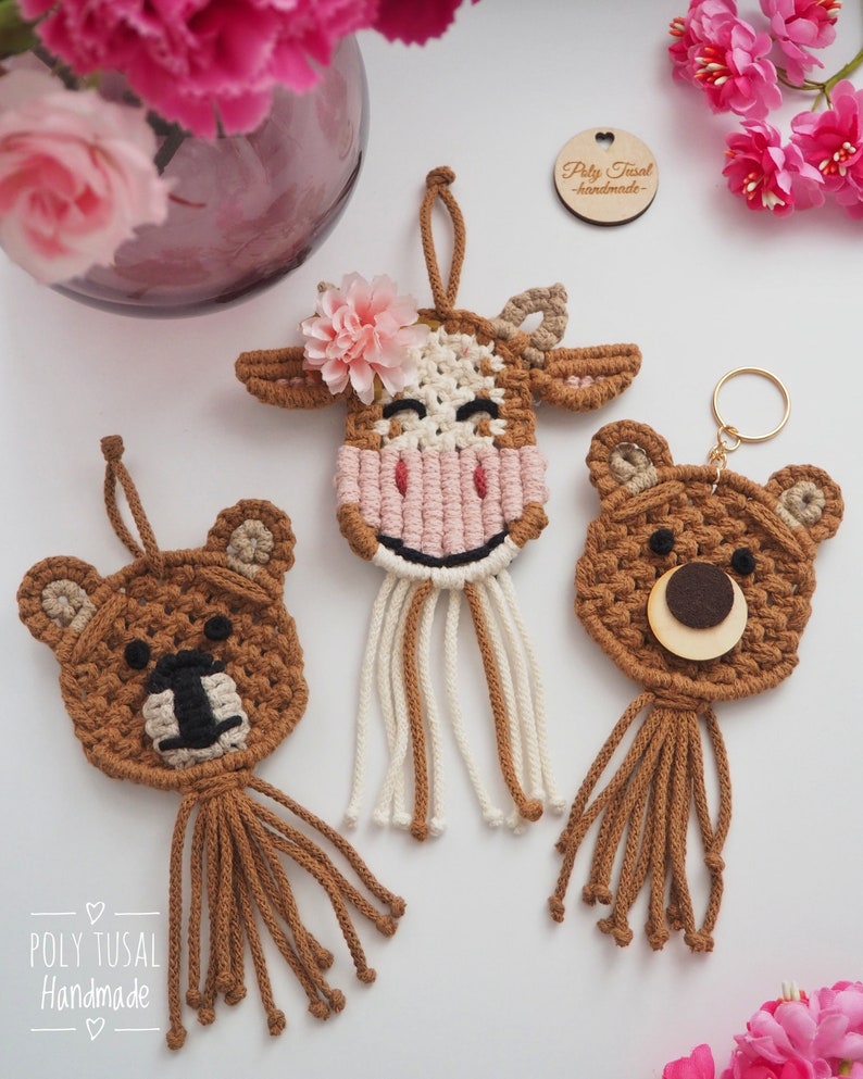 May include: Three handmade macrame animal keychains: a cow with a pink flower, and two bears. The cow has a pink and white face, brown ears, and a brown body. The bears are brown with black and wooden accents. The keychains have hanging loops and tassels.