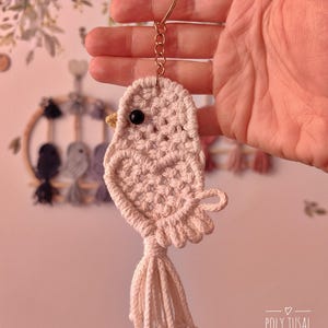 May include: A peach-coloured macrame bird keyring. The bird features a black bead eye, a yellow beak, and a tassel tail. It is suspended from a gold-coloured keyring and chain. The text "POLY TUSAL Handmade" is visible in the lower right corner.