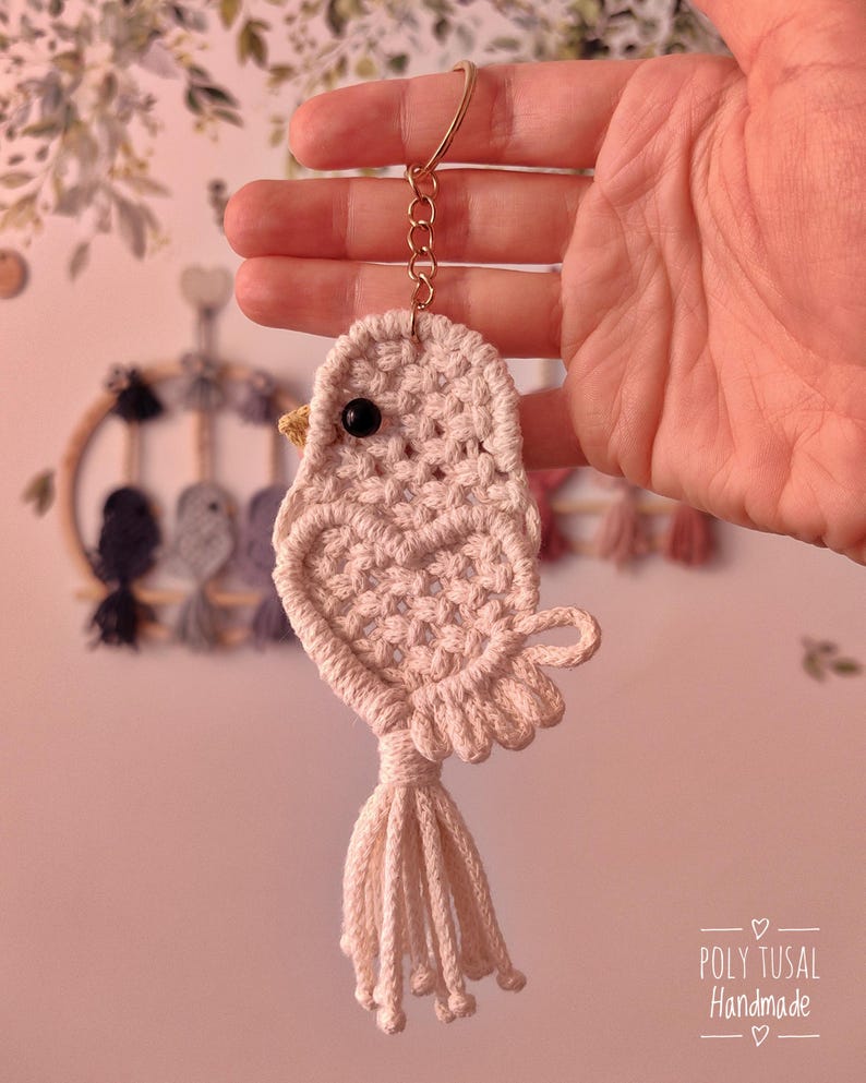 May include: A peach-coloured macrame bird keyring. The bird features a black bead eye, a yellow beak, and a tassel tail. It is suspended from a gold-coloured keyring and chain. The text "POLY TUSAL Handmade" is visible in the lower right corner.