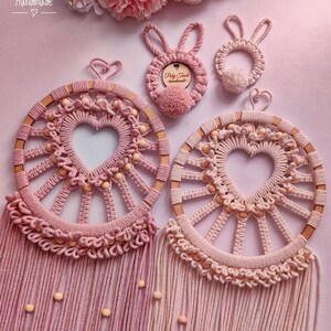 May include: Two pink macrame dreamcatchers with wooden beads and heart-shaped centers. The dreamcatchers have fringe and are hanging from a light pink background. There are two smaller pink macrame bunny ornaments with wooden beads and a tag that says "Poly Tusai Handmade".