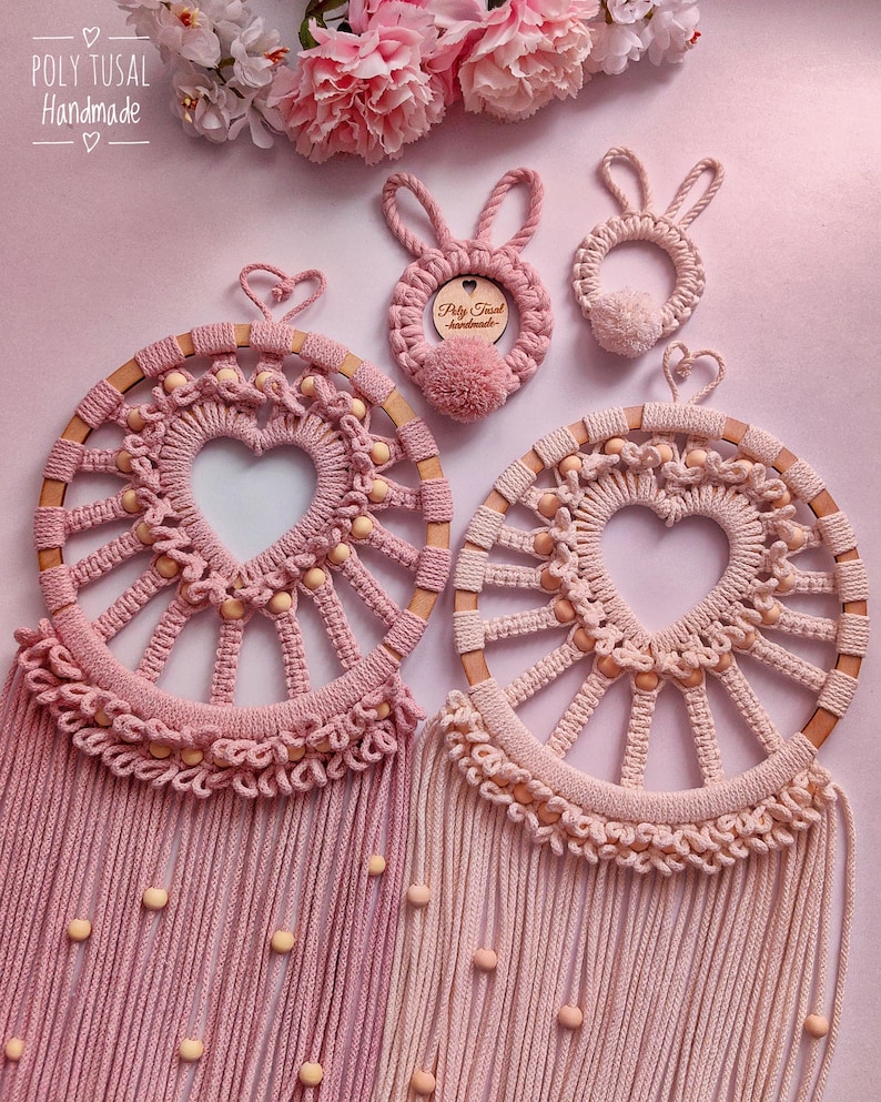 May include: Two pink macrame dreamcatchers with wooden beads and heart-shaped centers. The dreamcatchers have fringe and are hanging from a light pink background. There are two smaller pink macrame bunny ornaments with wooden beads and a tag that says "Poly Tusai Handmade".