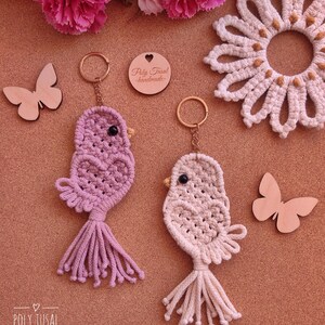 May include: Two macrame bird keyrings in lilac and cream, with gold rings. The birds have black bead eyes and a tassel tail. Wooden butterfly cutouts and a crochet flower are also visible.
