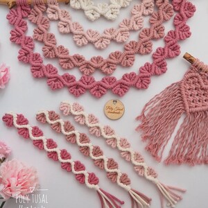 Op de afbeelding: Een set van drie roze en witte macram&eacute; wandhangers met hartvormen. De hangers zijn gemaakt van katoenen koord en hebben een franje aan de onderkant. De grootste hanger is bevestigd aan een tak. De kleinere hangers zijn in een rij onder de grootste hanger gerangschikt. De tekst "Poly Tusai Handmade" is zichtbaar in de linkerbenedenhoek van de afbeelding.