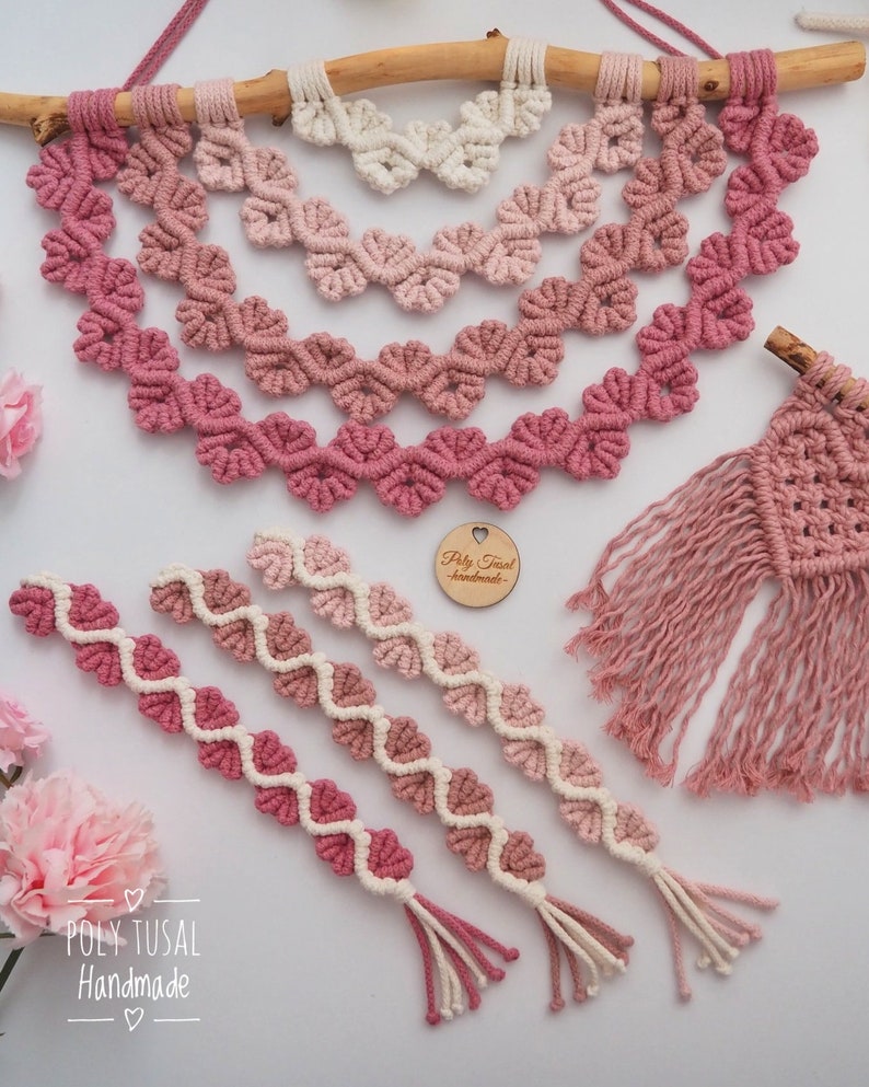 Op de afbeelding: Een set van drie roze en witte macram&eacute; wandhangers met hartvormen. De hangers zijn gemaakt van katoenen koord en hebben een franje aan de onderkant. De grootste hanger is bevestigd aan een tak. De kleinere hangers zijn in een rij onder de grootste hanger gerangschikt. De tekst "Poly Tusai Handmade" is zichtbaar in de linkerbenedenhoek van de afbeelding.
