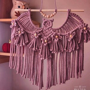 Pode incluir: Uma tape&ccedil;aria de parede de macram&eacute; feita &agrave; m&atilde;o em rosa empoeirado, com um design de coruja. A pe&ccedil;a &eacute; adornada com detalhes em forma de flor e contas de madeira. O design inclui longas franjas. O texto "POLY TUSAL Handmade" &eacute; vis&iacute;vel.