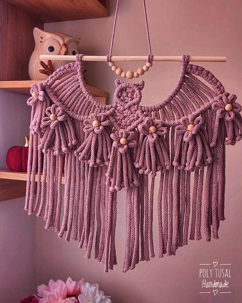 Pode incluir: Uma tape&ccedil;aria de parede de macram&eacute; feita &agrave; m&atilde;o em rosa empoeirado, com um design de coruja. A pe&ccedil;a &eacute; adornada com detalhes em forma de flor e contas de madeira. O design inclui longas franjas. O texto "POLY TUSAL Handmade" &eacute; vis&iacute;vel.
