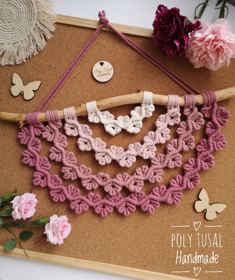 Pink version of Macrame Leaf Half Mandala laying on the cork board with pink flowers.  Next to Mandala are wooden butterflies.
On the right edge is logo of Poly Tusal Handmade.