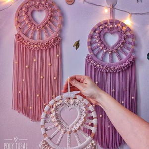 May include: Three handmade macrame dreamcatchers with wooden hoops and heart-shaped designs. The dreamcatchers are in pink, purple, and white with wooden beads.