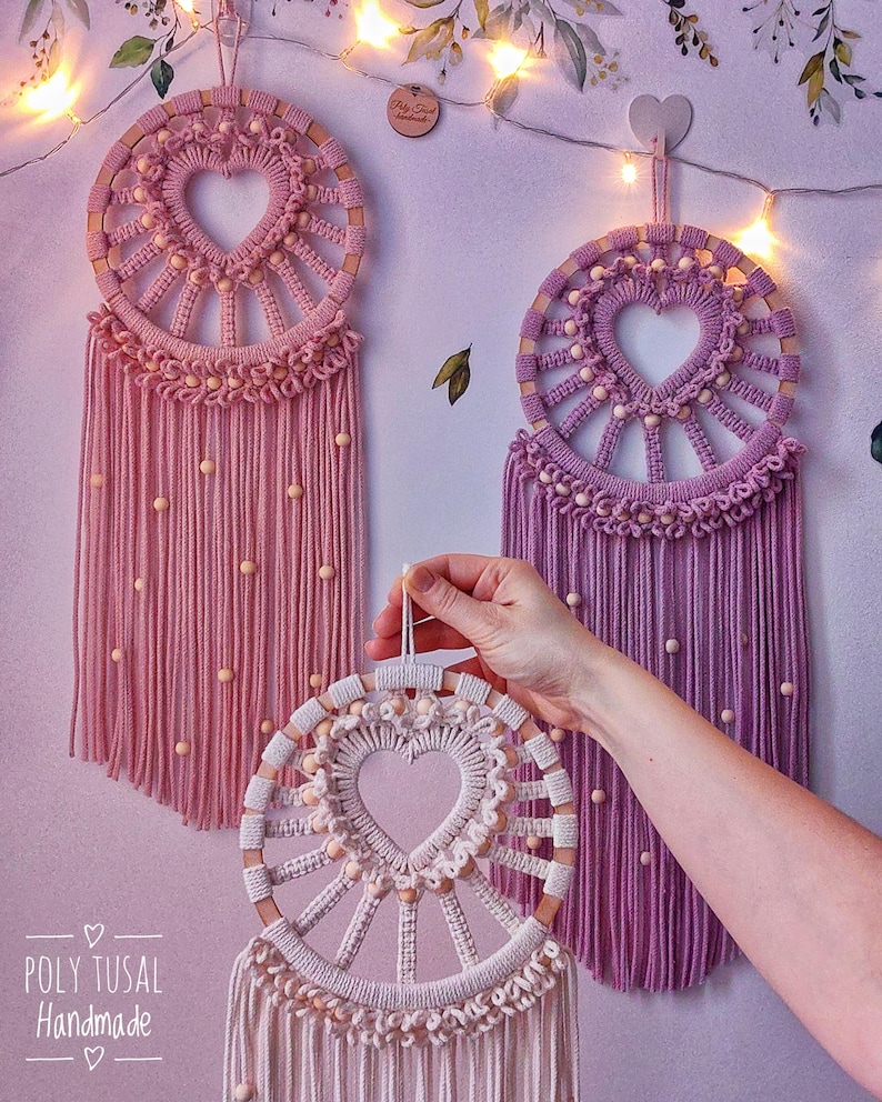 May include: Three handmade macrame dreamcatchers with wooden hoops and heart-shaped designs. The dreamcatchers are in pink, purple, and white with wooden beads.