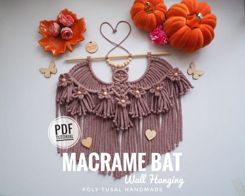 Pode incluir: Um pendurador de parede de morcego de macram&ecirc; rosa empoeirado com franjas e detalhes de flores de madeira. O texto "MACRAME BAT Wall Hanging" &eacute; exibido. Ab&oacute;boras decorativas, folhas e borboletas de madeira tamb&eacute;m est&atilde;o presentes.