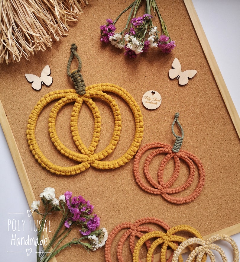 May include: Three macrame pumpkin wall hangings in different sizes. The largest pumpkin is yellow, the middle pumpkin is orange, and the smallest pumpkin is beige. The pumpkins are made with thick rope and have a rustic, handmade look. The pumpkins are hanging on a corkboard with a small wooden butterfly and a heart-shaped tag that says "Poly Tusul Handmade".