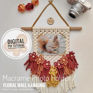 Könnte beinhalten: Ein Makramee-Bilderhalter mit floralem Design. Der Halter besteht aus einem weißen Seil mit roten, orangen und gelben Quasten. Ein kleines Holzschild mit dem Text "Poly Tusal Handmade" ist oben am Halter befestigt. Der Halter hängt an einer Holzleiste.