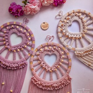 May include: Three handmade macrame dreamcatchers with wooden hoops and heart-shaped designs. The dreamcatchers are in shades of pink and white with wooden beads. The dreamcatchers are hanging from a white background with pink flowers.