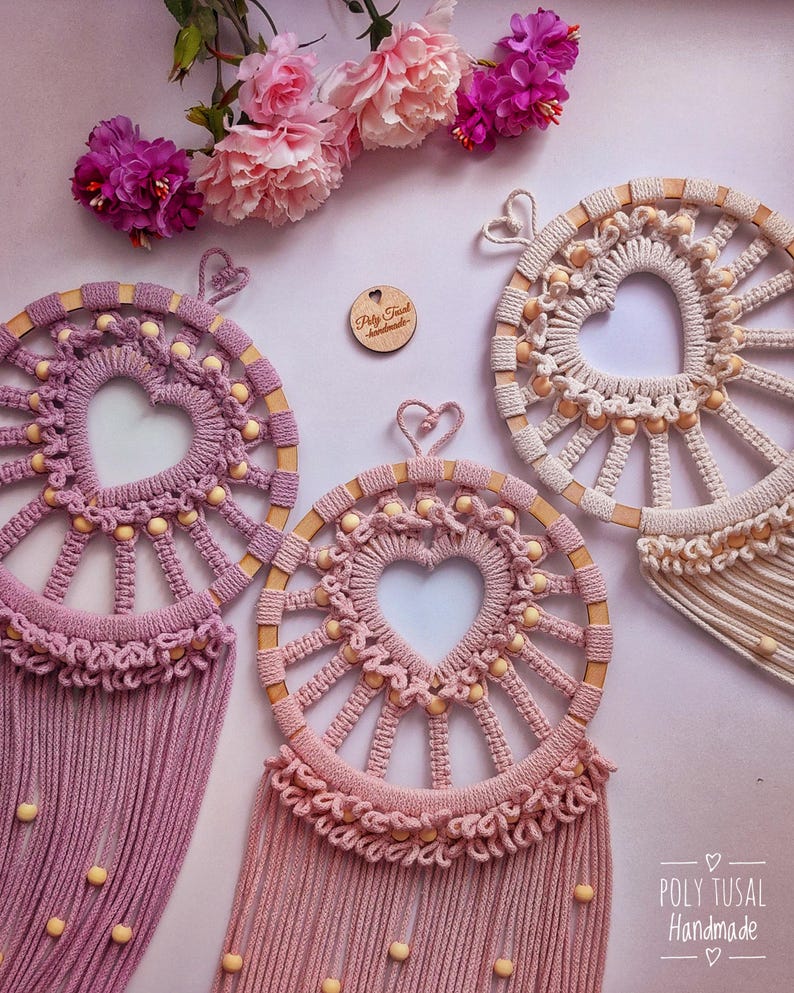 May include: Three handmade macrame dreamcatchers with wooden hoops and heart-shaped designs. The dreamcatchers are in shades of pink and white with wooden beads. The dreamcatchers are hanging from a white background with pink flowers.