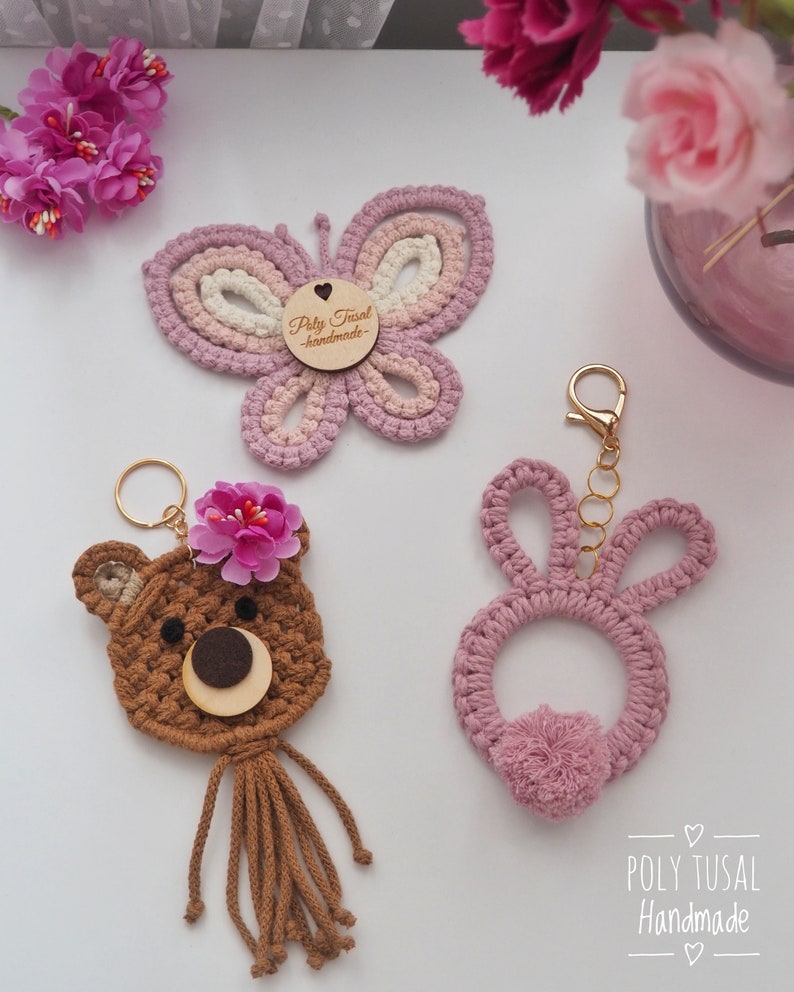 May include: A collection of handmade macrame keychains and decor. The image features a butterfly, a bear, and a bunny, all in shades of pink and brown. The butterfly has a wooden tag. The bear has a flower on its head. The bunny has a pom-pom tail.