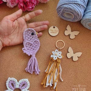 May include: A selection of handmade macrame keyrings and decorative items. The keyrings feature a purple bird and a white and yellow flower design. Other items include a white and purple flower, and wooden butterfly cutouts. The text "Poly Tusal - handmade" is visible.