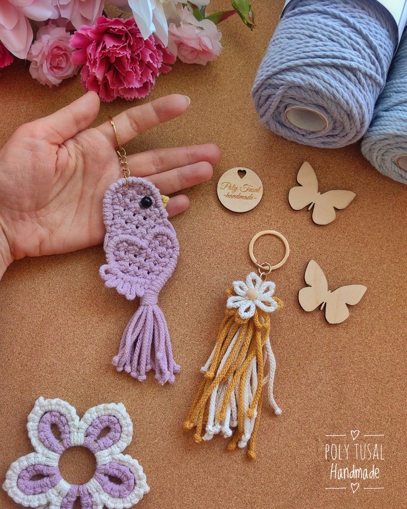 May include: A selection of handmade macrame keyrings and decorative items. The keyrings feature a purple bird and a white and yellow flower design. Other items include a white and purple flower, and wooden butterfly cutouts. The text "Poly Tusal - handmade" is visible.