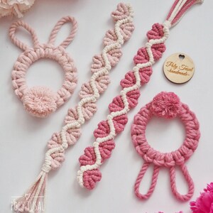 Op de afbeelding: Vier handgemaakte macram&eacute;-artikelen in roze en wit. Twee zijn rond met een pompon, &eacute;&eacute;n is een lange streng met een kwast en &eacute;&eacute;n is een lange streng met een hartvormig houten label waarop "Poly Tusai Handmade" staat.