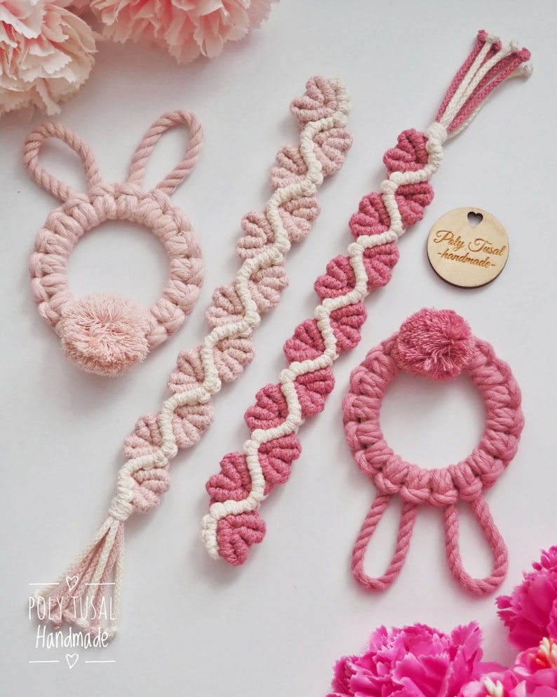 Op de afbeelding: Vier handgemaakte macram&eacute;-artikelen in roze en wit. Twee zijn rond met een pompon, &eacute;&eacute;n is een lange streng met een kwast en &eacute;&eacute;n is een lange streng met een hartvormig houten label waarop "Poly Tusai Handmade" staat.
