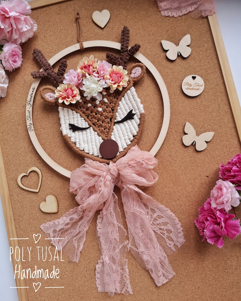 Cute sleeiing macrame deer with flower wreath on the forehead. The head is attaching to the plywood hoop. Over the neck is attaching a pink lace ribbon. Pink flowers in the edges of the photo.
