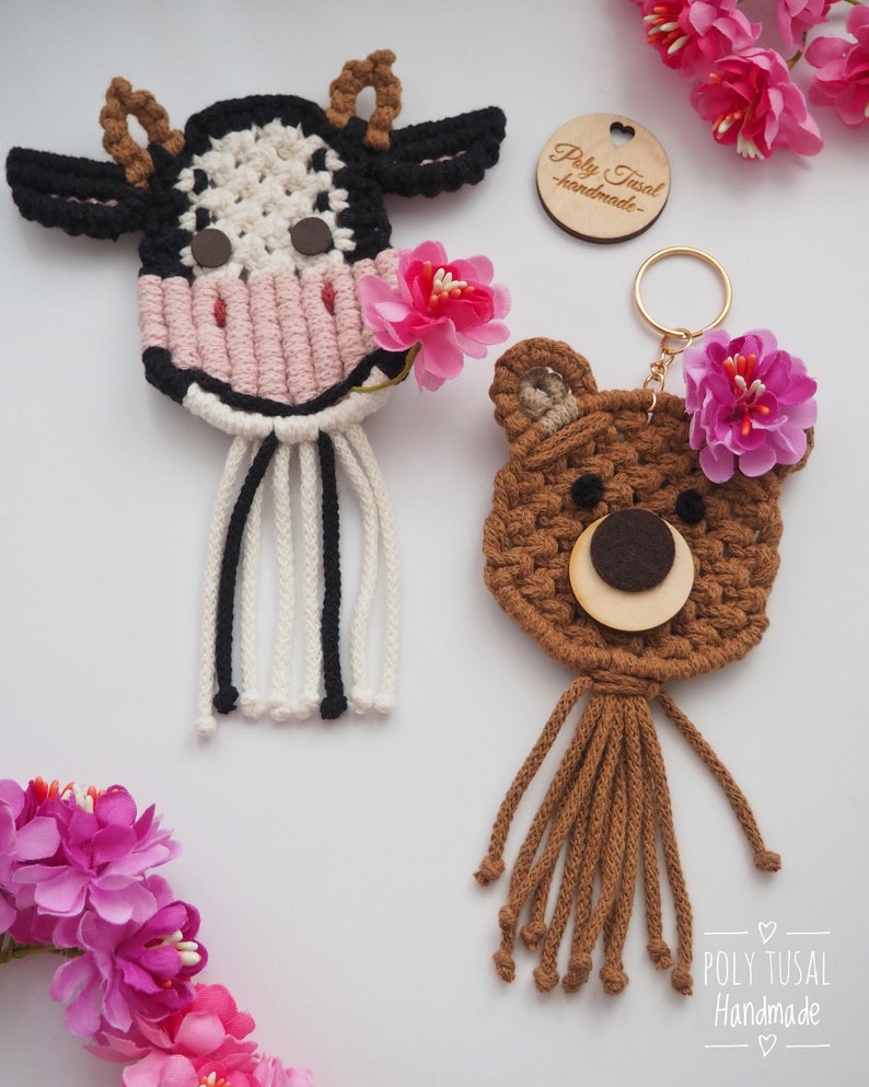 May include: Two handmade macrame keychains: a cow and a bear. The cow keychain features black and white details with a pink nose and a pink flower. The bear keychain is brown with a wooden face and a pink flower. Both have dangling cords.