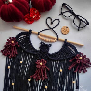 Pode incluir: Um pendurador de parede de macram&ecirc; feito &agrave; m&atilde;o em azul marinho, com borlas decorativas bord&ocirc; e contas de madeira. O design inclui um pino de madeira para pendurar. A imagem tamb&eacute;m mostra ab&oacute;boras de veludo vermelho, flores vermelhas e &oacute;culos pretos.