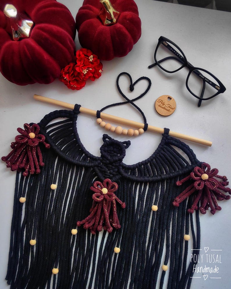 Pode incluir: Um pendurador de parede de macram&ecirc; feito &agrave; m&atilde;o em azul marinho, com borlas decorativas bord&ocirc; e contas de madeira. O design inclui um pino de madeira para pendurar. A imagem tamb&eacute;m mostra ab&oacute;boras de veludo vermelho, flores vermelhas e &oacute;culos pretos.