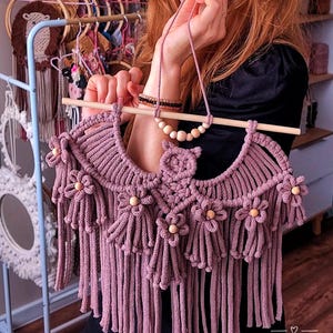 Pode incluir: Uma tape&ccedil;aria de parede de macram&eacute; rosa empoeirado feita &agrave; m&atilde;o, com franjas e detalhes de flores de madeira. A pe&ccedil;a &eacute; suspensa por um bast&atilde;o de madeira e apresenta um colar de contas a condizer. O texto "POLY TUSAL Handmade" &eacute; vis&iacute;vel.