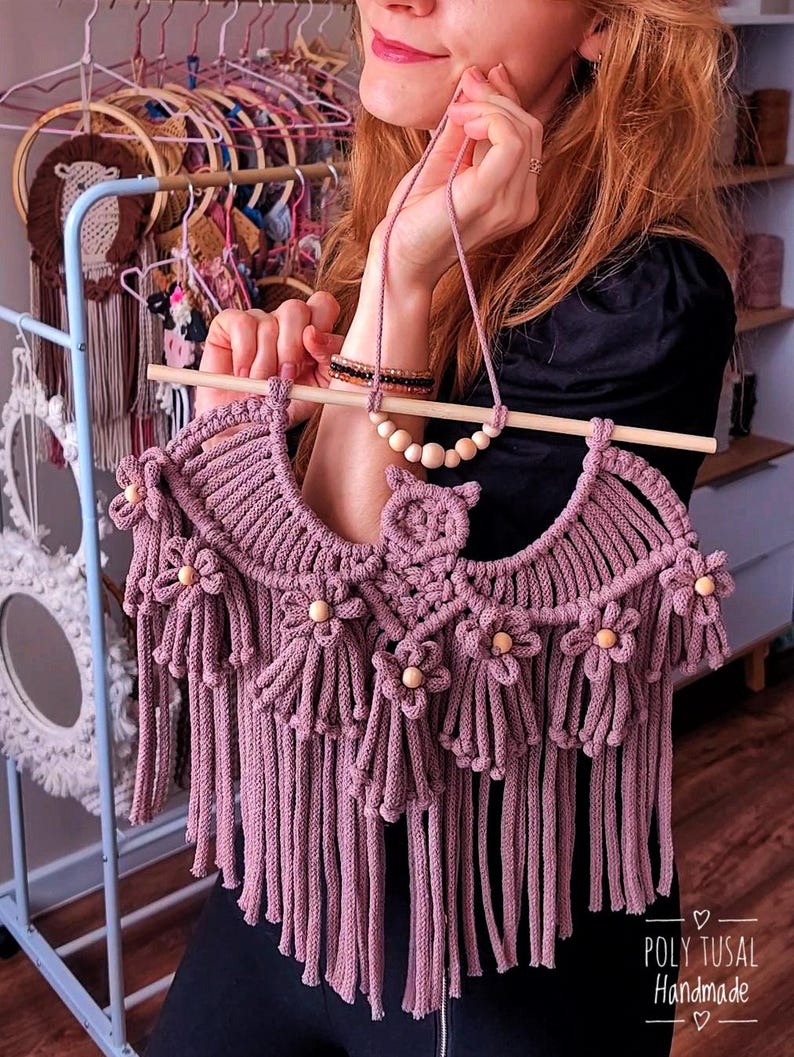 Pode incluir: Uma tape&ccedil;aria de parede de macram&eacute; rosa empoeirado feita &agrave; m&atilde;o, com franjas e detalhes de flores de madeira. A pe&ccedil;a &eacute; suspensa por um bast&atilde;o de madeira e apresenta um colar de contas a condizer. O texto "POLY TUSAL Handmade" &eacute; vis&iacute;vel.