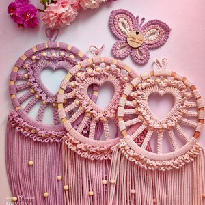 May include: Three pink and purple macrame dreamcatchers with wooden beads and a heart-shaped design. The dreamcatchers are hanging from a pink background. The text "Poly Tusai Handmade" is visible in the bottom left corner.