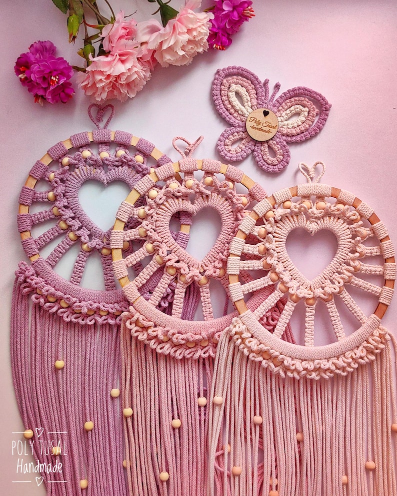 May include: Three pink and purple macrame dreamcatchers with wooden beads and a heart-shaped design. The dreamcatchers are hanging from a pink background. The text "Poly Tusai Handmade" is visible in the bottom left corner.