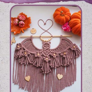 Pode incluir: Um pendurador de parede de morcego de macram&ecirc; feito &agrave; m&atilde;o em rosa empoeirado, com um design de franjas e detalhes de cora&ccedil;&atilde;o de madeira. A decora&ccedil;&atilde;o de parede inclui um pino de madeira e &eacute; adornada com pequenos detalhes florais. O texto "MACRAME BAT WALL HANGING" &eacute; vis&iacute;vel.