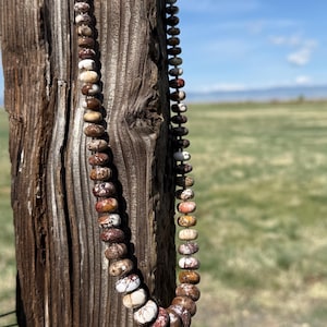 May include: A beaded necklace with brown, white, and reddish-brown stones hangs against a weathered wooden post. The necklace features a variety of rounded beads, showcasing natural color variations. The background includes a field and a blue sky.