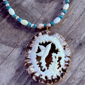 Pode incluir: Um colar artesanal com um pingente esculpido em um material castanho claro, apresentando um design de beija-flor. O colar inclui contas turquesas e brancas, juntamente com espaçadores prateados, criando um acessório inspirado na natureza.