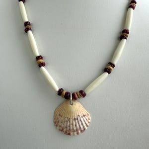 May include: A handmade necklace with a seashell pendant. The necklace features cream-coloured, elongated beads with small, dark red and brown beads. The seashell pendant is light tan with reddish-purple accents.