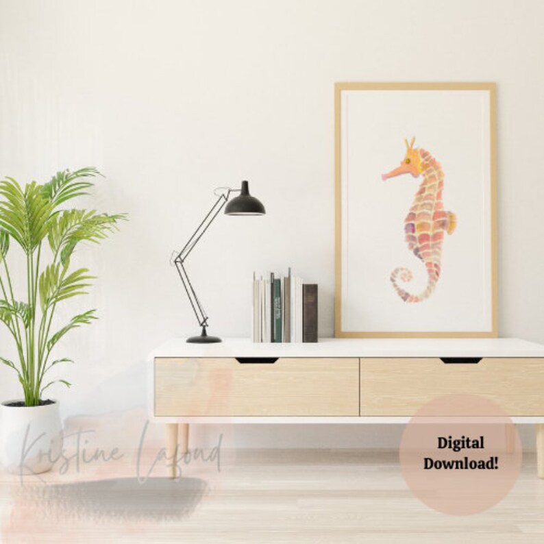 Puede incluir: Una impresi&oacute;n en acuarela enmarcada de un caballito de mar en tonos naranja, rosa y morado. El caballito de mar es el punto focal, sobre un fondo blanco dentro de un marco de madera clara. La impresi&oacute;n se exhibe en un mueble blanco y de madera.