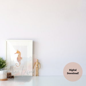 Puede incluir: Una impresi&oacute;n en acuarela enmarcada de un caballito de mar en tonos naranja y rosa. La obra de arte se muestra en un marco blanco y descansa sobre una superficie blanca. El texto "Kristine Lafond" es visible en la impresi&oacute;n. Un maniqu&iacute; de artista de madera y una peque&ntilde;a planta est&aacute;n cerca.