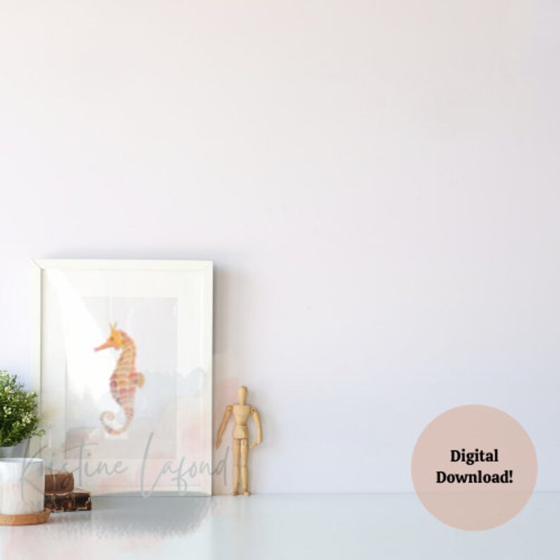 Puede incluir: Una impresi&oacute;n en acuarela enmarcada de un caballito de mar en tonos naranja y rosa. La obra de arte se muestra en un marco blanco y descansa sobre una superficie blanca. El texto "Kristine Lafond" es visible en la impresi&oacute;n. Un maniqu&iacute; de artista de madera y una peque&ntilde;a planta est&aacute;n cerca.