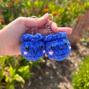 Puede incluir: Dos llaveros de peluche azul hechos a crochet con caras sonrientes y mejillas rosadas. Los llaveros están unidos a cadenas plateadas.