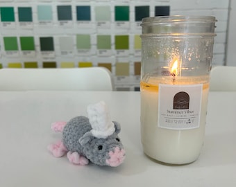 May include: A lit candle in a glass jar next to a crocheted gray mouse wearing a white top hat. The candle has a label that reads "Summer Vibes". The mouse has pink feet and nose.