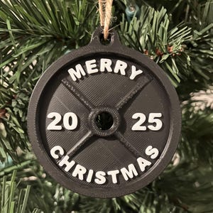 May include: A black weight plate Christmas ornament with the words "MERRY CHRISTMAS" in white. The numbers "20" and "25" are also in white. The ornament is hanging from a natural twine string. The ornament is hanging in front of a green Christmas tree.