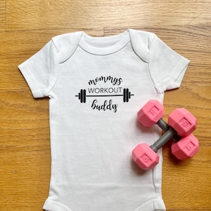 May include: White baby onesie with the text "mommy's workout buddy" in black. The onesie is next to two pink dumbbells. The dumbbells have a gray handle and are marked with the number 5.